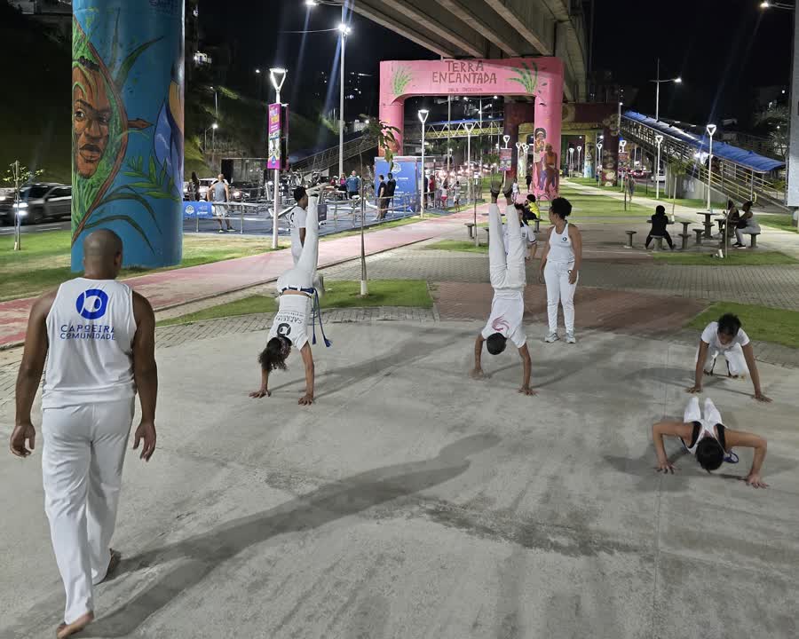 Treino acontecendo à noite no canteiro central, sob o viaduto: várias pessoas de branco em bananeira sobre o piso de concreto, a coluna com o mural pintado à esquerda e o arco cor-de-rosa da Terra Encantada ao fundo.