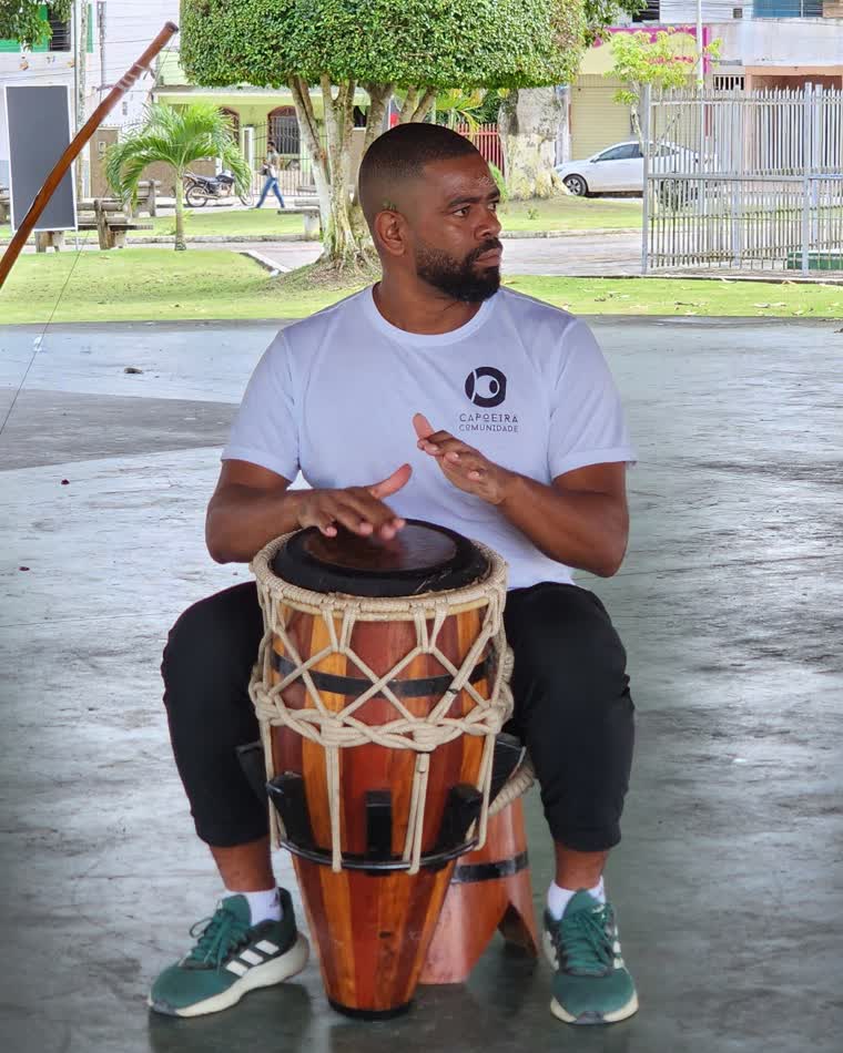 Contramestre Queimadinho tocando atabaque, de camisa do Capoeira Comunidade.