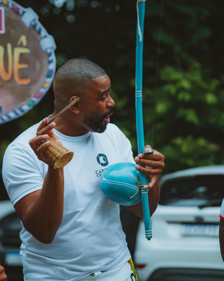 Tocando berimbau, com o caxixi na mão e a camisa do grupo.