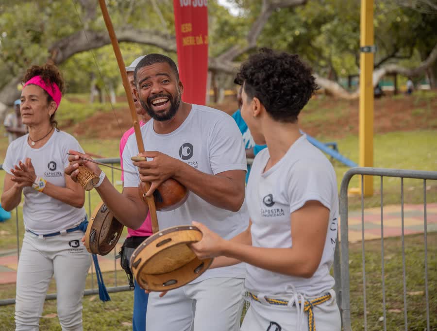 Três integrantes do grupo rindo enquanto tocam: uma mulher de bandana rosa batendo palma, um homem com o berimbau e uma jovem com o pandeiro.